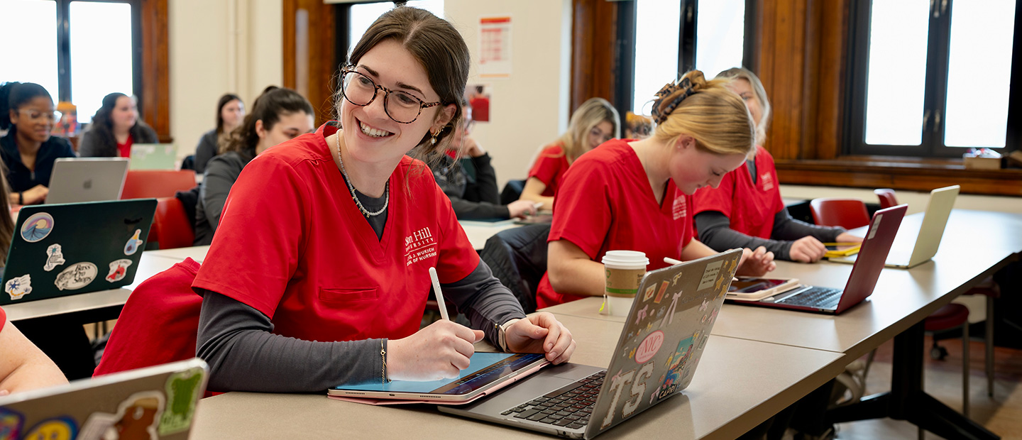 Nursing students wearing red scrubs work on laptops and tablets in a classroom.