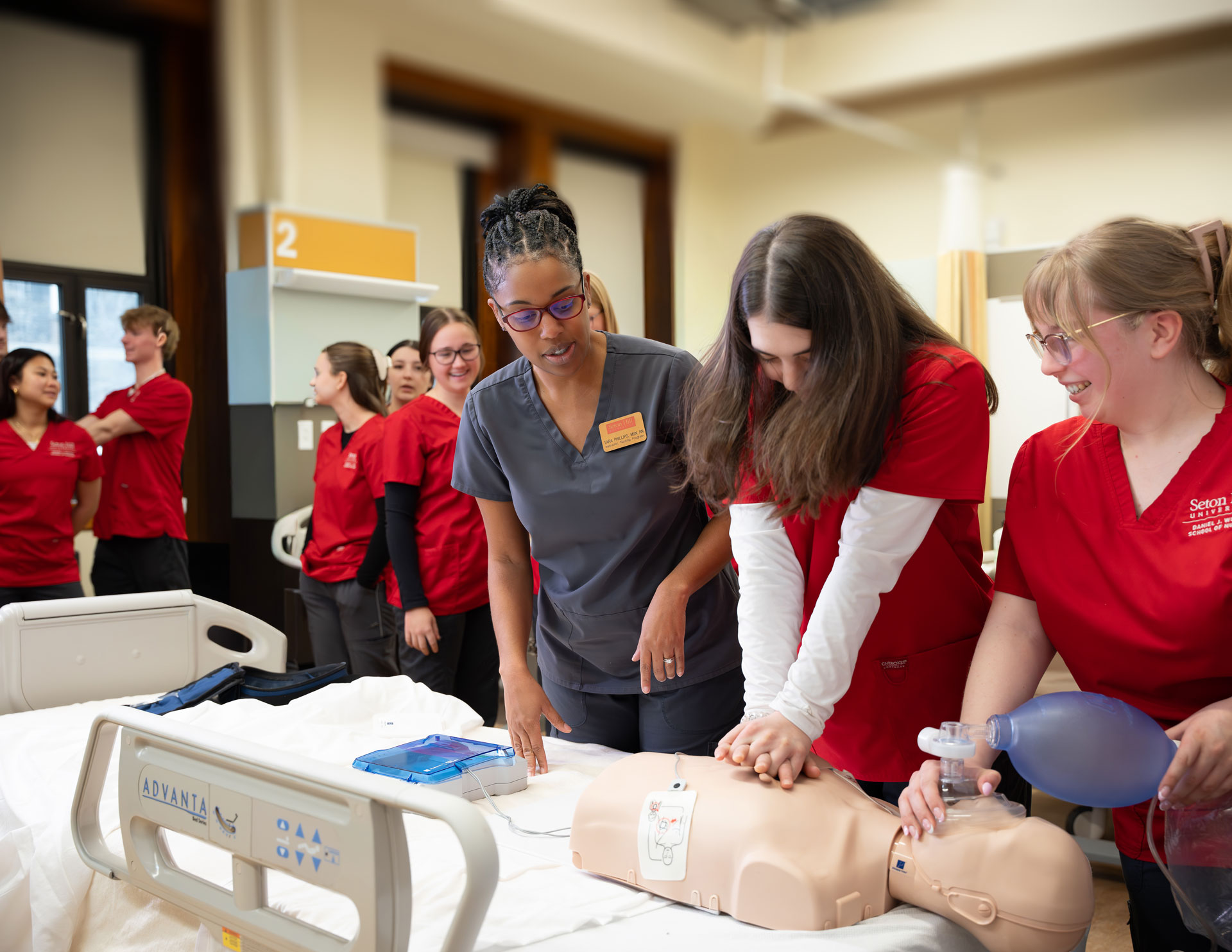 A group of nursing students in red scrubs work with their instructor in a simulation lab.